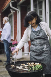 Glückliche Frau mit Schürze grillt Gemüse auf einem Grill auf einer Party - MASF32226