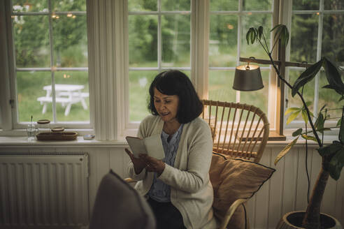 Reife Frau, die ihr Smartphone benutzt, während sie auf einem Stuhl in der Nähe des Fensters zu Hause sitzt - MASF32249