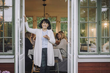 Porträt einer Frau mit Strickjacke, die ein Glas in der Hand hält und an der Tür steht - MASF32286