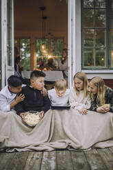Excited friends with blanket and popcorn watching tablet PC while sitting on porch - MASF32291