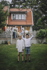 Playful boys with soccer ball standing in back yard with family on porch in background - MASF32296