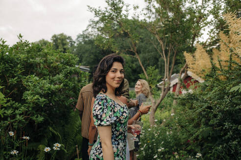 Side view of smiling woman with drink standing by plants in garden - MASF32312