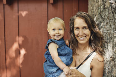 Porträt einer glücklichen Mutter, die ihre Tochter trägt und sich auf einen Baumstamm stützt - MASF32351