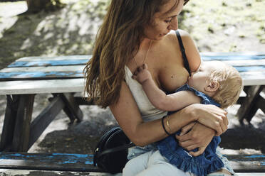 Mother feeding breast milk to toddler daughter while sitting on bench - MASF32353