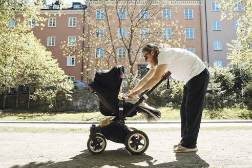 Side view of man playing with daughter in baby stroller while standing on footpath - MASF32356