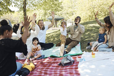Glückliche Eltern mit erhobenen Armen sitzen mit Kindern auf einer Picknickdecke im Park - MASF32361