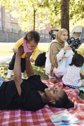 Woman playing with daughter while lying down on picnic blanket at park - MASF32365