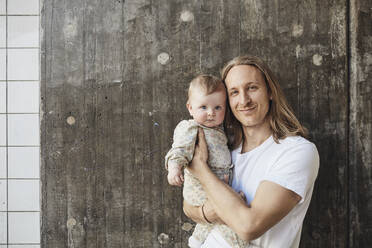 Porträt eines lächelnden Mannes mit langen Haaren, der seine kleine Tochter vor einer Wand trägt - MASF32374