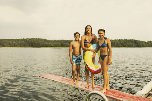 Lächelndes Mädchen mit aufblasbarem Ring steht mit Geschwistern auf einem Sprungbrett am See - MASF32386