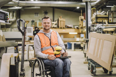 Portrait of mature carpenter with ear protectors and digital tablet sitting on wheelchair in warehouse - MASF32427