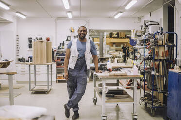 Porträt eines lächelnden Tischlers in voller Länge, der auf einem Bein steht und sich in der Werkstatt auf eine Werkbank stützt - MASF32431