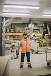 Full length of smiling carpenter wearing reflective clothing and ear protectors in warehouse - MASF32436