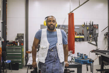 Porträt eines lächelnden Tischlers mit Gehörschutz, der sich in einer Werkstatt an eine Werkbank lehnt - MASF32442