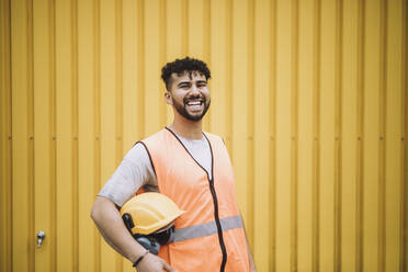 Portrait of cheerful young construction worker with hardhat standing against yellow metal wall - MASF32461