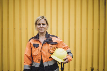 Porträt einer reifen Bauarbeiterin mit Schutzhelm vor einer gelben Metallwand - MASF32462