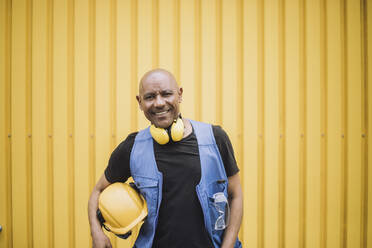 Portrait of happy bald construction worker with hardhat and ear protectors in front of yellow metal wall - MASF32465
