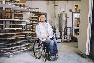 Full length of smiling painter wearing ear protectors while sitting on wheelchair in workshop - MASF32484