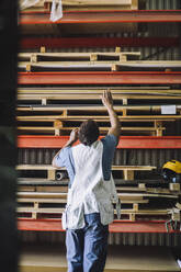 Rear view of male carpenter searching for wooden plank on pallet stack in warehouse - MASF32497