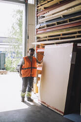 Full length of young carpenter in reflective clothing standing by wooden plank at warehouse - MASF32507