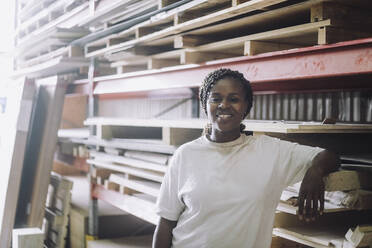 Portrait of happy carpenter standing by pallet stack in warehouse - MASF32522