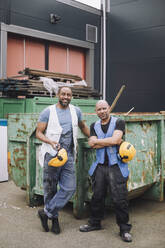 Männliche Bauarbeiter in voller Länge mit Schutzhelm vor einem Metallcontainer auf der Baustelle stehend - MASF32556