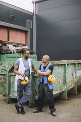 Lächelnde männliche Kollegen mit Schutzhelm, die sich gegenseitig gegen einen Metallcontainer ansehen - MASF32557