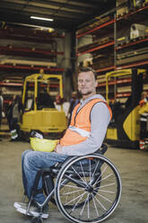 Side view of carpenter with hardhat sitting on wheelchair in warehouse - MASF32585