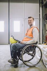 Side view of carpenter with hardhat and digital tablet sitting on wheelchair in warehouse - MASF32599