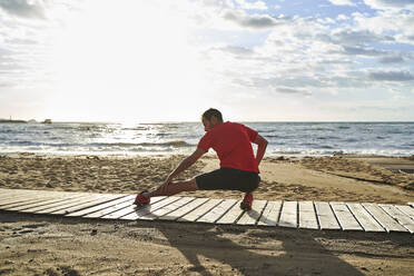Man stretching leg on boardwalk at beach - VEGF06063