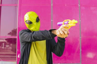 Man wearing alien mask playing with toy gun in front of pink wall - MGRF00821