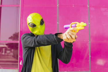 Mann mit Alien-Maske spielt mit Spielzeugpistole vor einer rosa Wand - MGRF00821