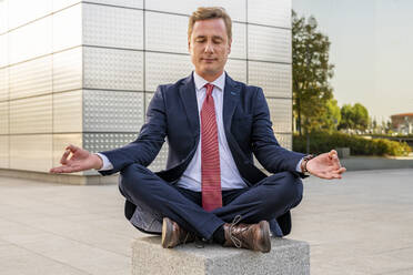 Businessman in lotus position meditating on concrete block - DLTSF03147