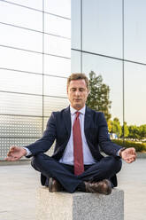 Mature businessman in lotus position meditating on concrete block - DLTSF03148