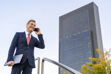 Happy mature businessman holding laptop talking on mobile phone by railing - DLTSF03150