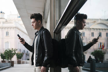 Junger Mann mit Smartphone an einer Glaswand lehnend - EGAF02584
