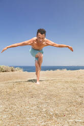 Shirtless man practicing eagle pose yoga on field in front of sea on sunny day - ACTF00292