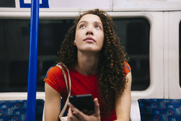 Nachdenkliche junge Frau mit Smartphone in der U-Bahn sitzend - AMWF00899