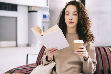 Young woman reading book with disposable cup of coffee - AMWF00918