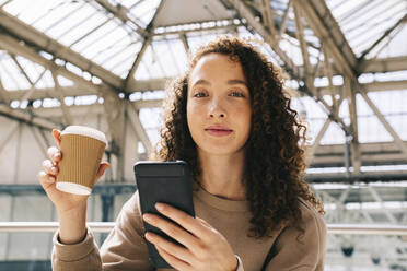 Smiling woman sitting with mobile phone drinking coffee at train station - AMWF00923