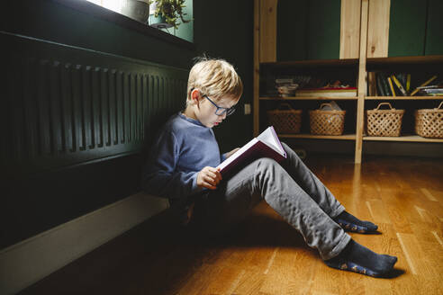 Junge liest Buch auf dem Boden sitzend zu Hause - EYAF02230