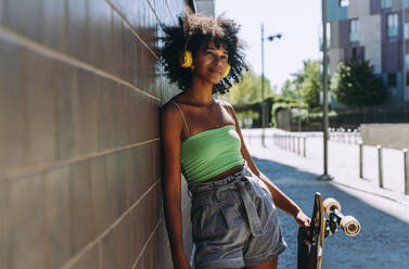 Smiling young woman wearing headphones with skateboard leaning on wall - OIPF02418