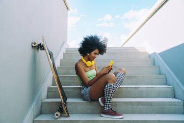 Woman using mobile phone sitting by skateboard on staircase - OIPF02446