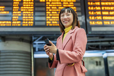 Smiling young woman standing with smart phone in front of time board - DLTSF03175