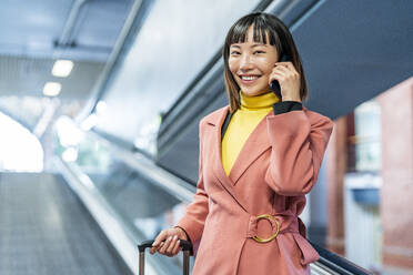 Glückliche junge Frau, die mit einem Smartphone spricht und einen Koffer auf einer Rolltreppe hält - DLTSF03176