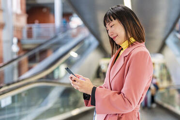 Lächelnde junge Frau, die auf einer Rolltreppe eine Textnachricht über ihr Smartphone sendet - DLTSF03179