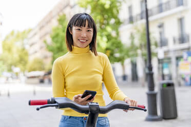 Glückliche junge Frau mit Smartphone auf einem Elektrofahrrad stehend - DLTSF03196