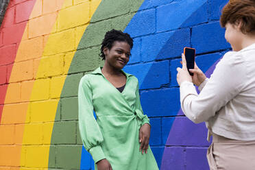 Lächelnde Frau, die ihre Freundin vor einer an die Wand gemalten Regenbogenflagge fotografiert - AMWF00931