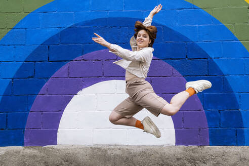 Smiling young woman with hand raised jumping in front of colorful wall - AMWF00940