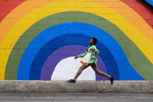 Smiling young woman holding mesh bag jumping in front of rainbow flag painted on wall - AMWF00943