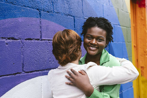 Smiling woman embracing friend by colorful wall - AMWF00950